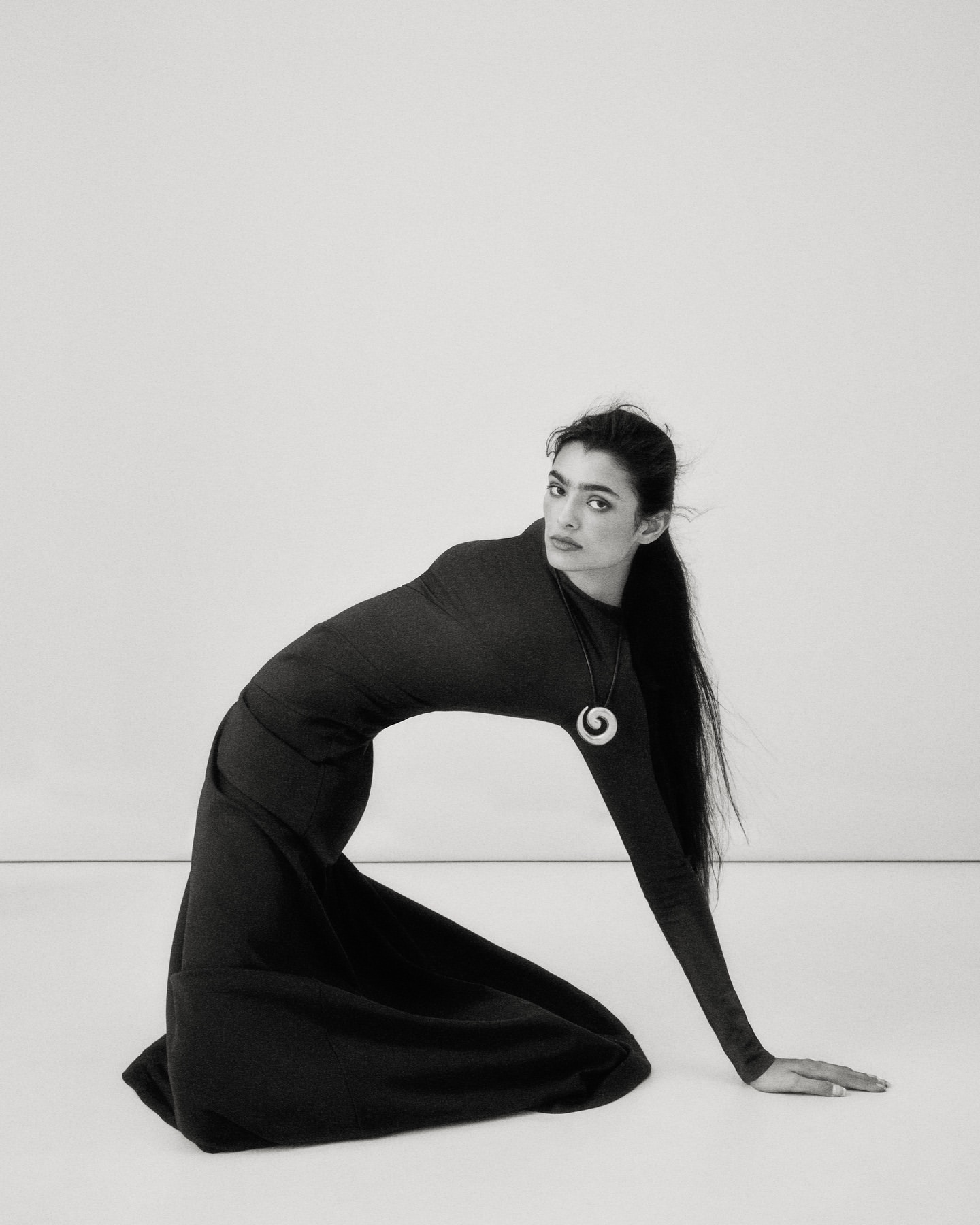 Monochrome portrait of a brunette woman in a black dress kneeling, featuring a simple white backdrop and eye-level camera angle.