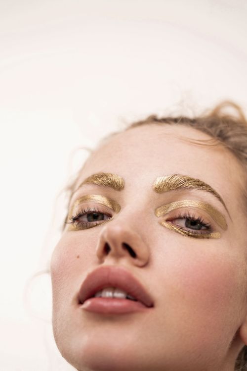 Close-up shot of a fair-skinned adult woman with golden makeup on her eyebrows and eyelids, against a bright, neutral background.