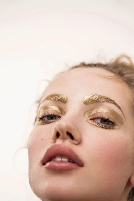 Close-up shot of a fair-skinned adult woman with golden makeup on her eyebrows and eyelids, against a bright, neutral background.