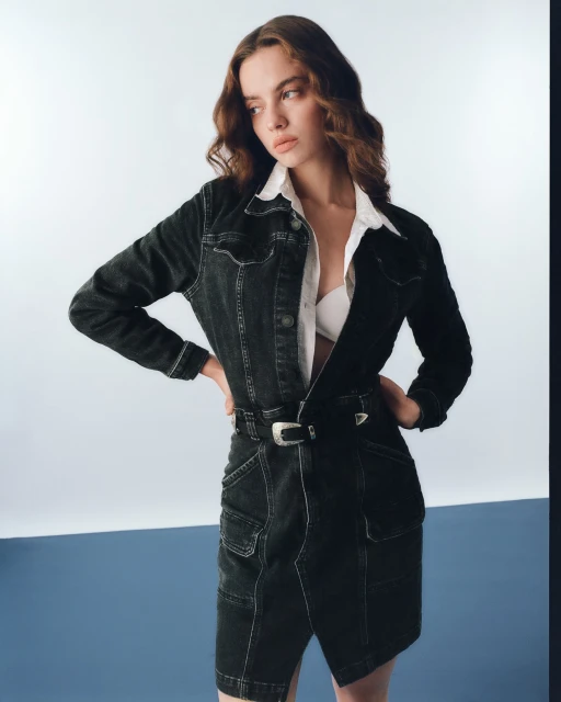 A young woman in a black denim jacket and skirt poses against a split blue and white backdrop, creating a modern, fashionable look.