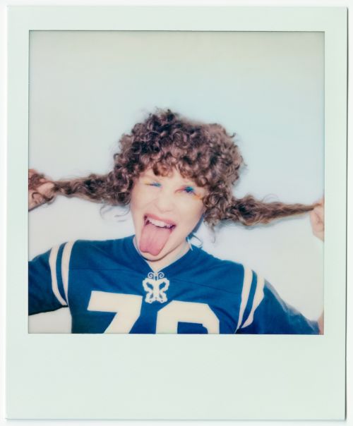 Young woman with curly hair sticks out her tongue while playfully pulling her hair in a photo booth-style portrait.