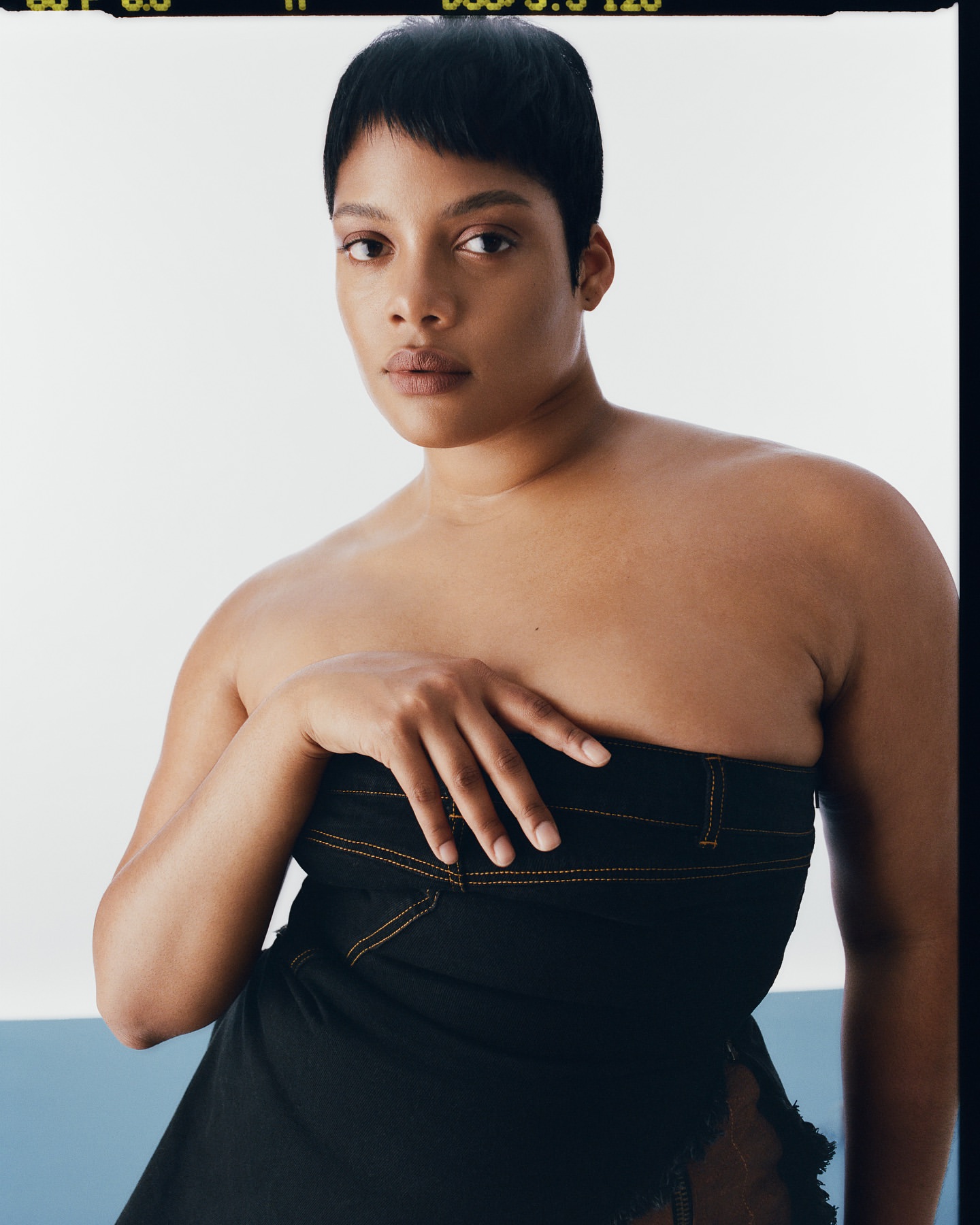 Close-up studio portrait of an adult woman with short dark hair wearing a strapless dark dress against a split white and light blue backdrop.