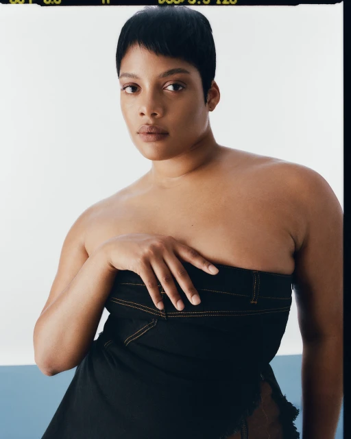 Close-up studio portrait of an adult woman with short dark hair wearing a strapless dark dress against a split white and light blue backdrop.