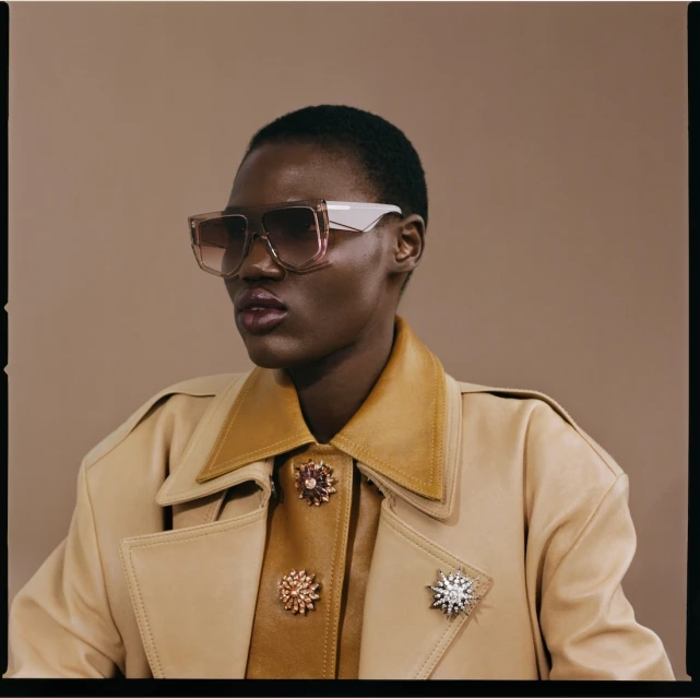 Elegant adult female wearing oversized sunglasses, tan trench coat with decorative brooches in a studio setting.