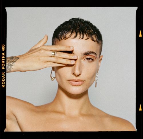 Portrait of woman with short hair covering eye with hand wearing gold earrings and a ring posing against a neutral background.