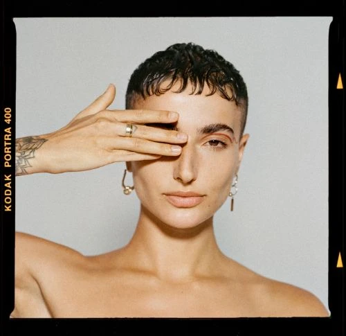 Portrait of woman with short hair covering eye with hand wearing gold earrings and a ring posing against a neutral background.