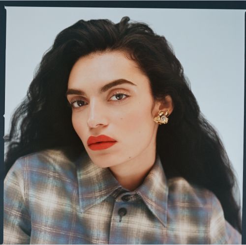 Portrait of a woman with dark wavy hair wearing a plaid shirt and gold earrings against a light background