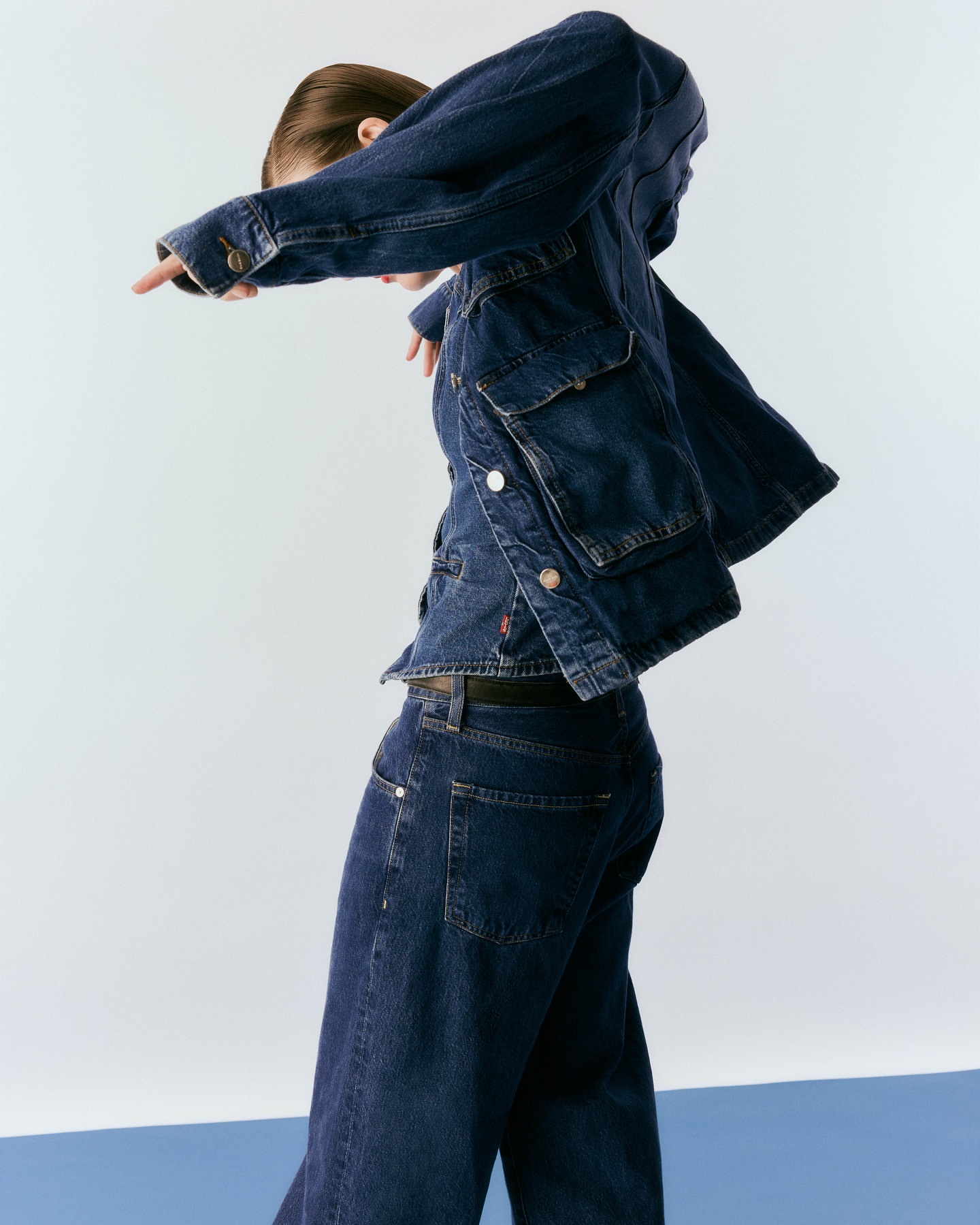 Adult female in stylish denim jacket and jeans strikes dynamic pose in a light photo studio featuring a blue and white background.
