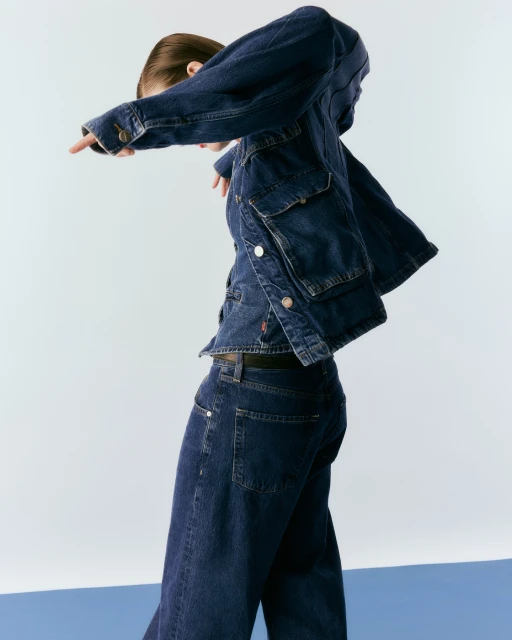 Adult female in stylish denim jacket and jeans strikes dynamic pose in a light photo studio featuring a blue and white background.