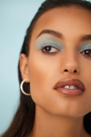Close-up portrait of a woman with blue eyeshadow and silver hoop earrings against a pale blue background