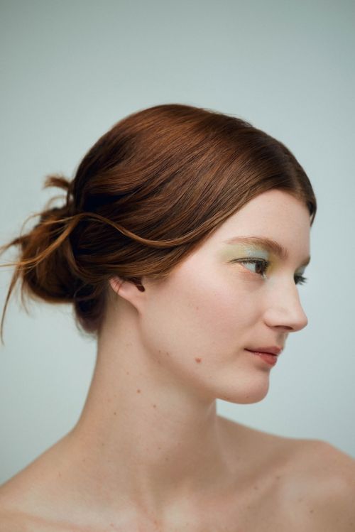 Close-up portrait of a woman with auburn hair styled in an updo and light green eyeshadow against a light blue backdrop.