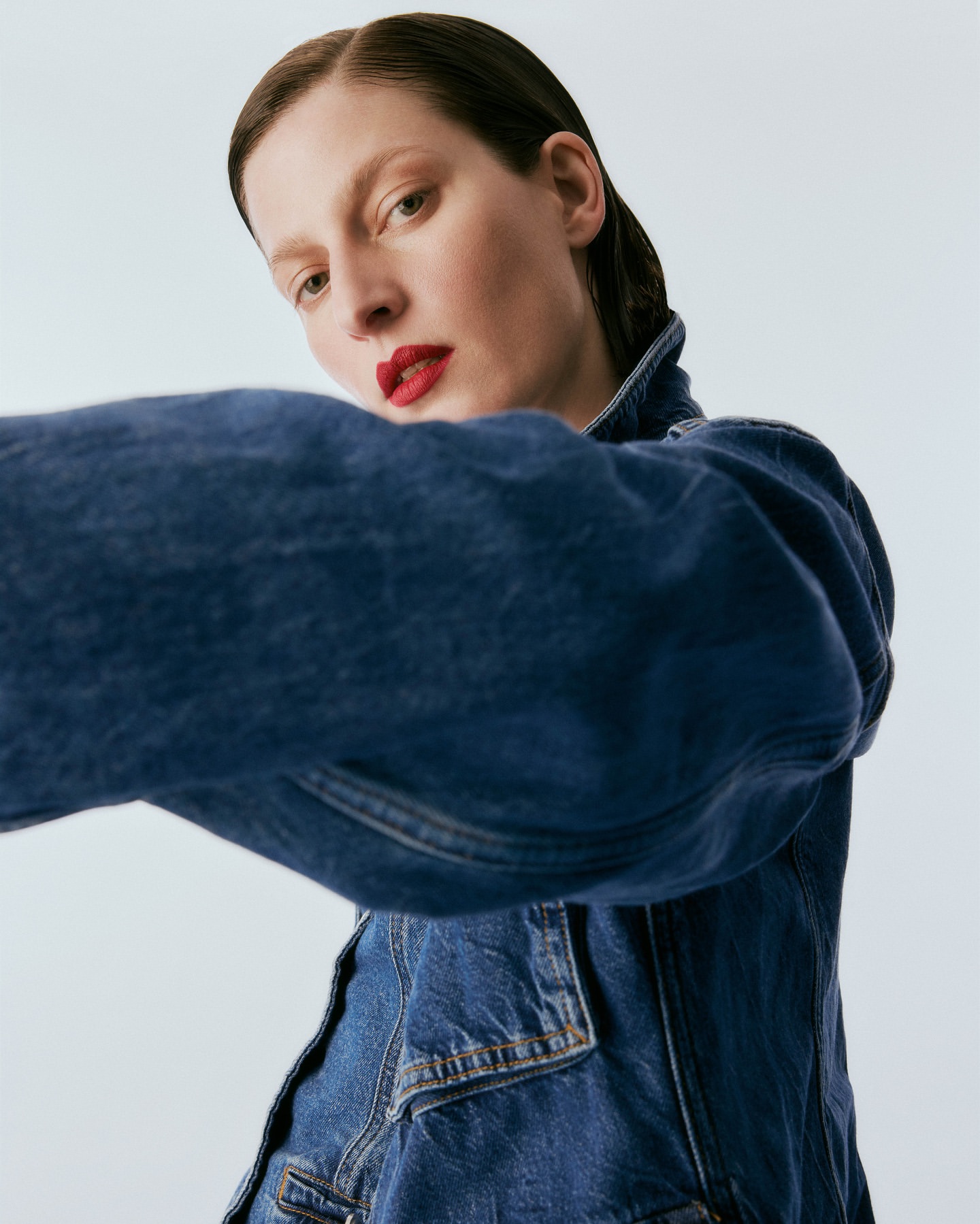 Model wearing a blue denim jacket with dark slicked-back hair and bold red lipstick against a plain white background