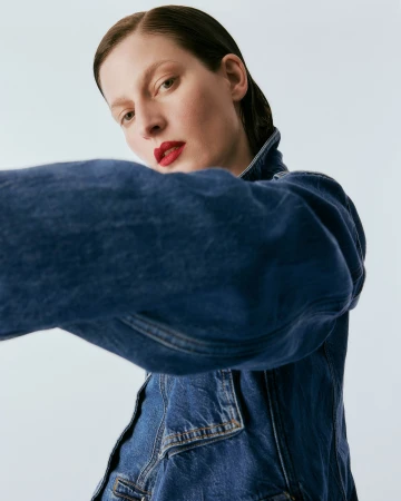 Model wearing a blue denim jacket with dark slicked-back hair and bold red lipstick against a plain white background