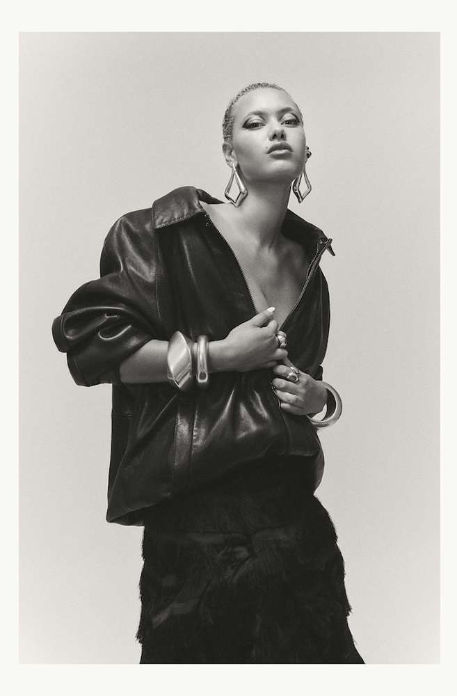 Fashion portrait of a young woman with platinum hair wearing a black leather jacket, large earrings, and bracelets against a light background.