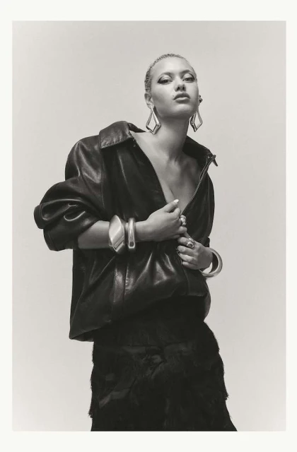 Fashion portrait of a young woman with platinum hair wearing a black leather jacket, large earrings, and bracelets against a light background.