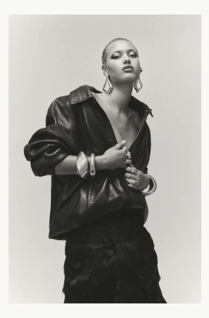 Fashion portrait of a young woman with platinum hair wearing a black leather jacket, large earrings, and bracelets against a light background.