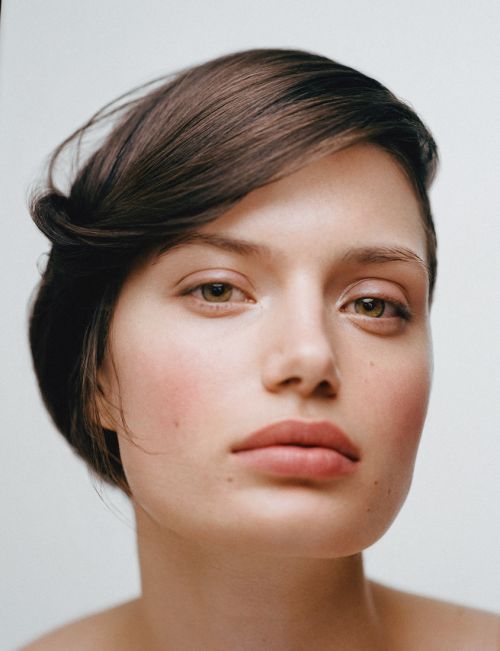 Close-up portrait of a young woman with fair skin, short brown hair, and brown eyes against a plain white background in a studio setting.