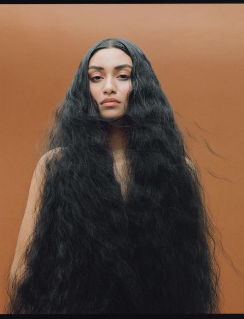Portrait of a woman with long, wavy black hair against an orange backdrop, looking directly at the camera with a neutral expression.