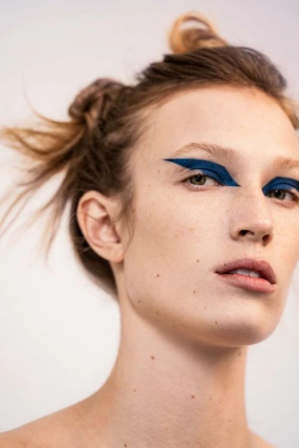 Close-up portrait of a woman with fair skin and auburn hair in small buns, posing against a white backdrop wearing bold blue eye makeup.