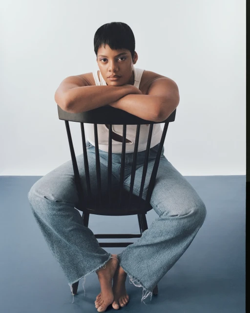 An adult woman with a short haircut is sitting backwards on a wooden chair in a studio. She is wearing a white top and jeans.