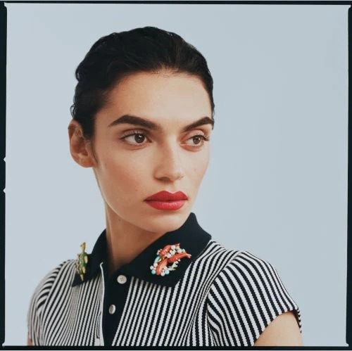 Portrait of a woman with dark hair and red lipstick wearing a striped polo shirt with decorative brooches against a light blue background.
