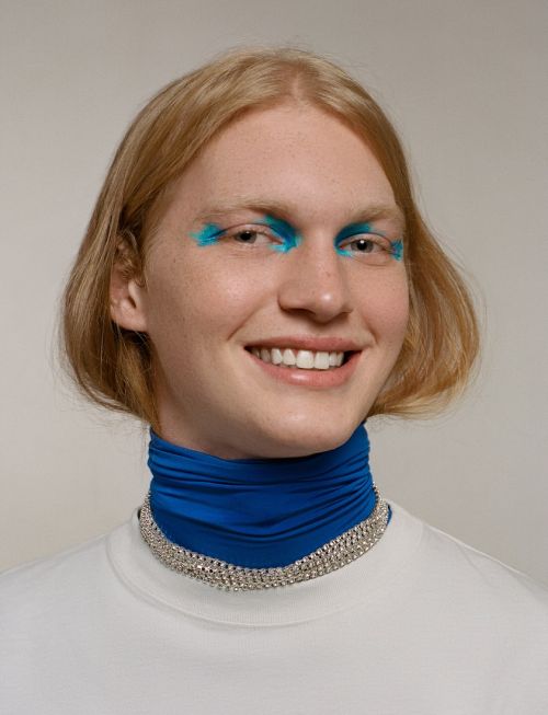Close-up of smiling young adult with blonde hair wearing blue eye makeup, white t-shirt, blue neck scarf, and silver choker necklace.