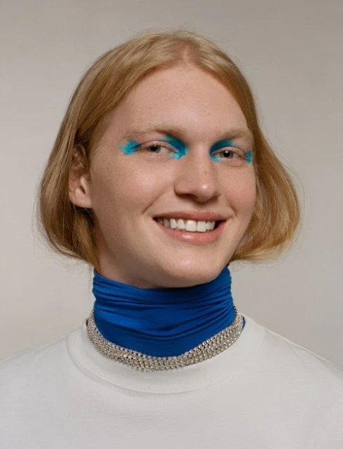Close-up of smiling young adult with blonde hair wearing blue eye makeup, white t-shirt, blue neck scarf, and silver choker necklace.