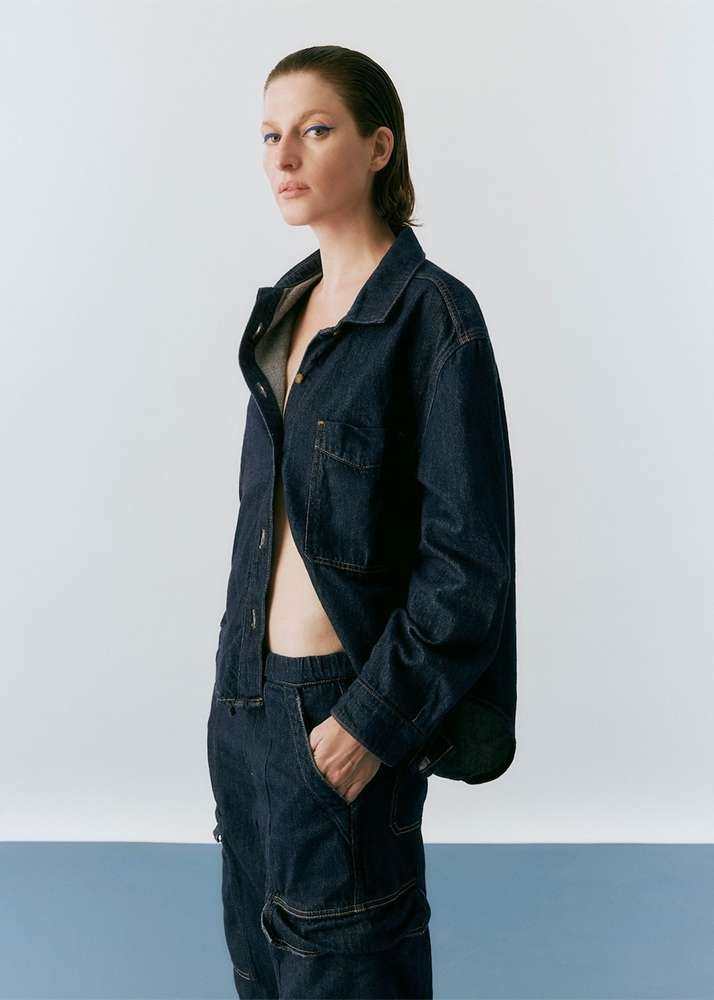 A woman with wet hair poses in a studio wearing a dark-denim jacket and matching pants with a pale-blue and white backdrop.