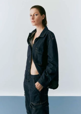 A woman with wet hair poses in a studio wearing a dark-denim jacket and matching pants with a pale-blue and white backdrop.
