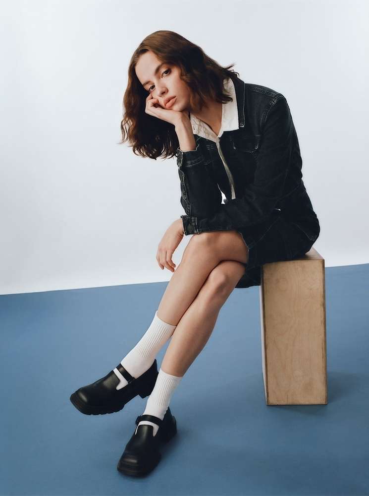 A young woman poses indoors wearing a dark denim jacket and skirt, white socks, and black Mary Jane shoes, seated on a wooden block.