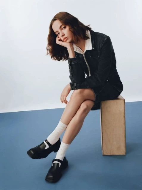 A young woman poses indoors wearing a dark denim jacket and skirt, white socks, and black Mary Jane shoes, seated on a wooden block.