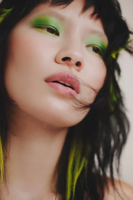 Close-up portrait of a woman with green eyeshadow and black hair with green streaks, looking up with soft lighting.