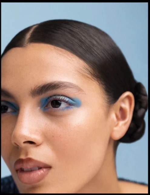 Close-up portrait of a woman with dark hair and bright blue eyeshadow against a light blue background in a beauty or fashion context.
