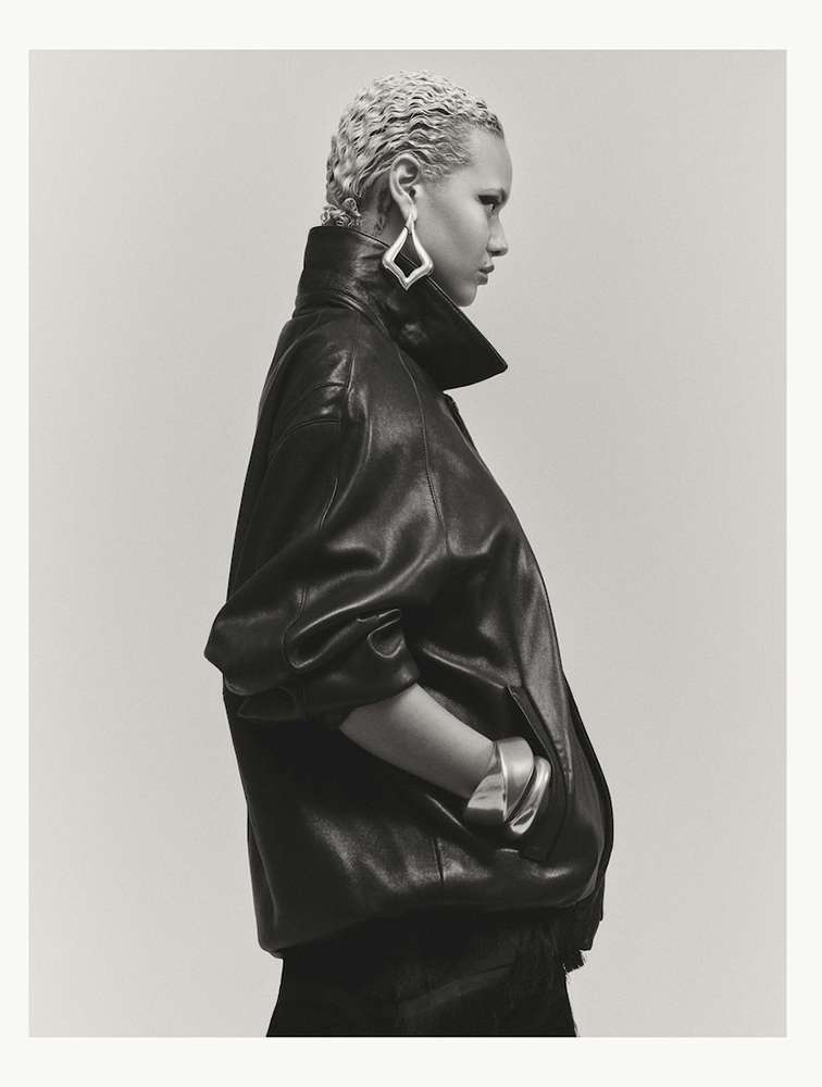 Model in dark leather jacket with light blonde braided hair wearing geometric silver earrings against a plain backdrop