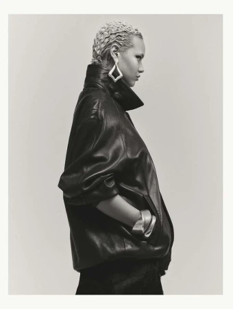 Model in dark leather jacket with light blonde braided hair wearing geometric silver earrings against a plain backdrop