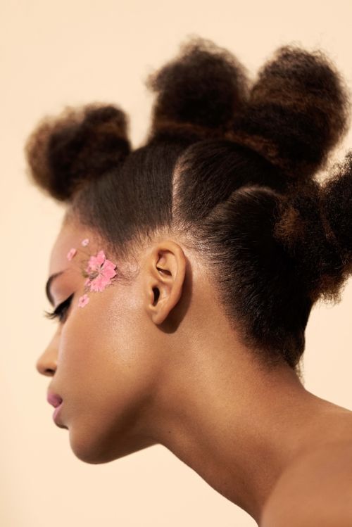 Profile portrait of a woman with pink makeup and flowers by her eye, styled afro hair, and a peach-toned background.