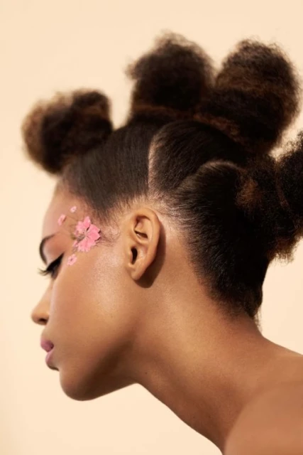 Profile portrait of a woman with pink makeup and flowers by her eye, styled afro hair, and a peach-toned background.