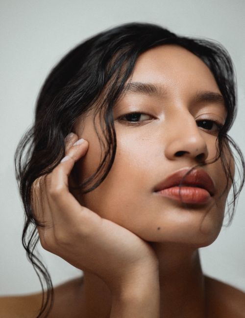 Close-up portrait of a woman with dark wavy hair and light skin, gazing directly at the camera with a neutral expression on a neutral background.