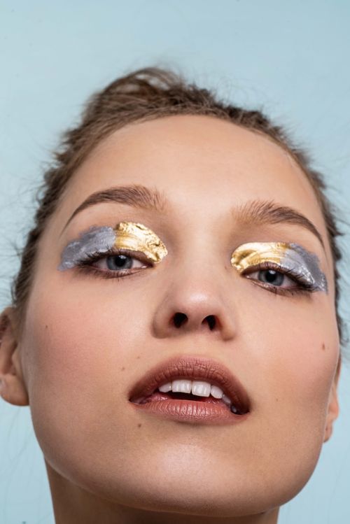 Close-up portrait of a woman with creative gold and silver foil makeup on her eyelids, posing against a light blue backdrop.