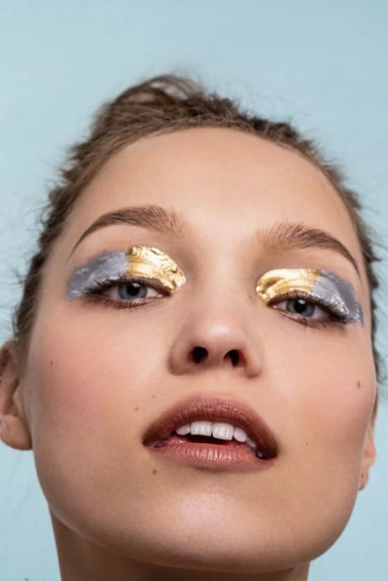 Close-up portrait of a woman with creative gold and silver foil makeup on her eyelids, posing against a light blue backdrop.