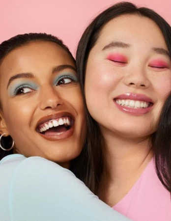 Close-up portrait of two smiling young women with colorful makeup embracing against a pink backdrop