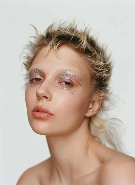 Close-up portrait of a young adult female with blonde hair, creative eye makeup, and pale skin against a white background in a beauty shot.