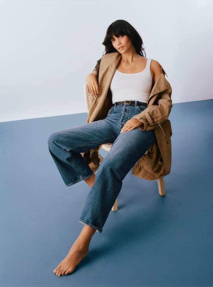Woman with dark hair seated on a wooden chair wearing a white tank top, blue jeans, and a draped camel coat against a blue and white studio backdrop.