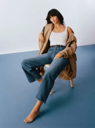 Woman with dark hair seated on a wooden chair wearing a white tank top, blue jeans, and a draped camel coat against a blue and white studio backdrop.