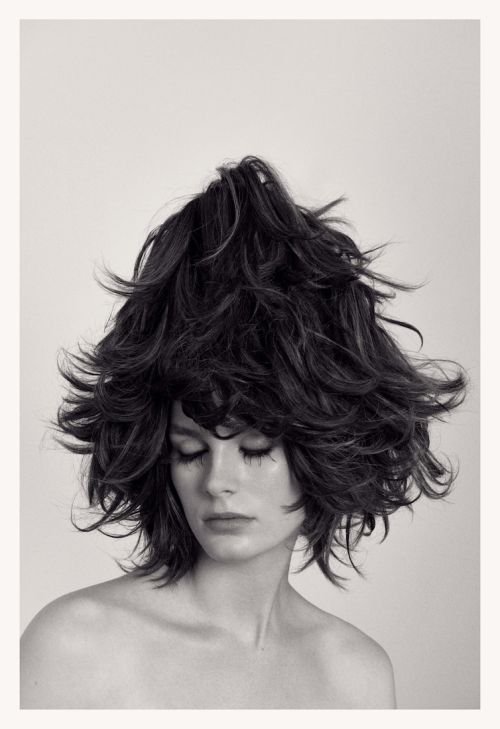 Headshot of woman with voluminous, dark, layered hair styled upwards, eyes closed, bare shoulders, against a light neutral background.