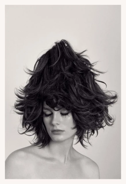 Headshot of woman with voluminous, dark, layered hair styled upwards, eyes closed, bare shoulders, against a light neutral background.