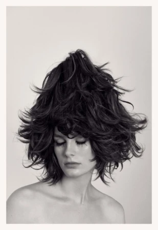 Headshot of woman with voluminous, dark, layered hair styled upwards, eyes closed, bare shoulders, against a light neutral background.