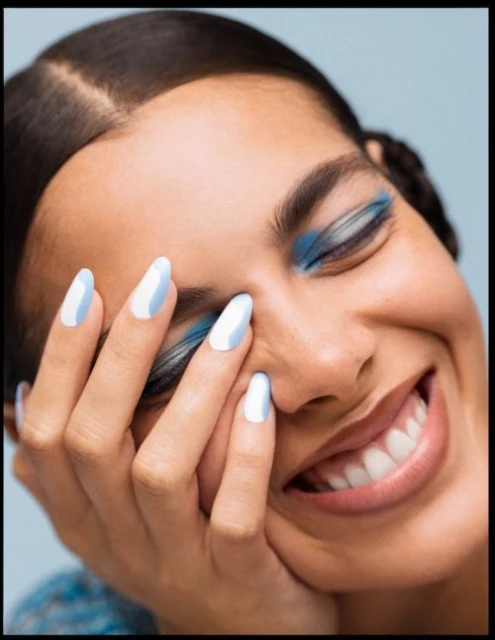 Close-up of a smiling woman with blue eyeshadow and blue and white nails, set against a light blue backdrop in a beauty shot.