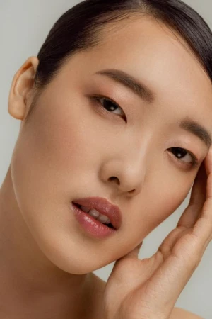 Close-up portrait of an East Asian woman with glossy pink lips and hand near face against a plain light background.
