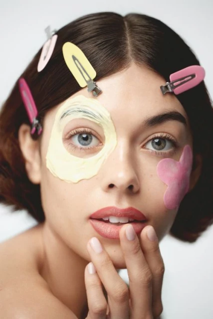 Close-up portrait of a young woman with skincare patches and hair clips, showcasing beauty and self-care in a light, neutral setting.